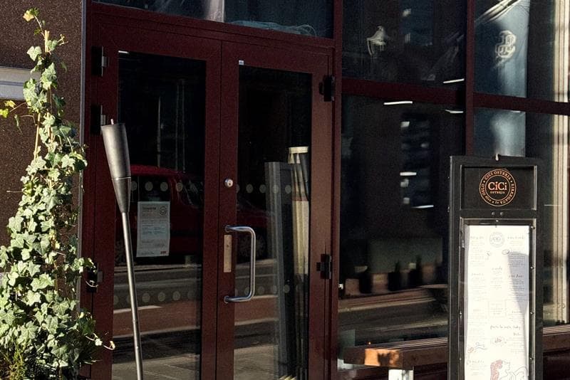 Inngangspartiet til en restaurant med store glassvinduer, menyplakat ved døren og en lykt og plante plassert på fortauet.